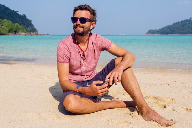 Happy attractive man in stylish sunglasses with beard using mobile phone at the beach