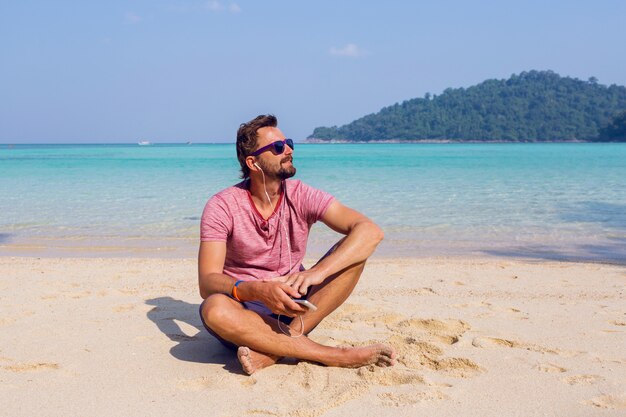 Happy attractive man in stylish sunglasses with beard using mobile phone at the beach