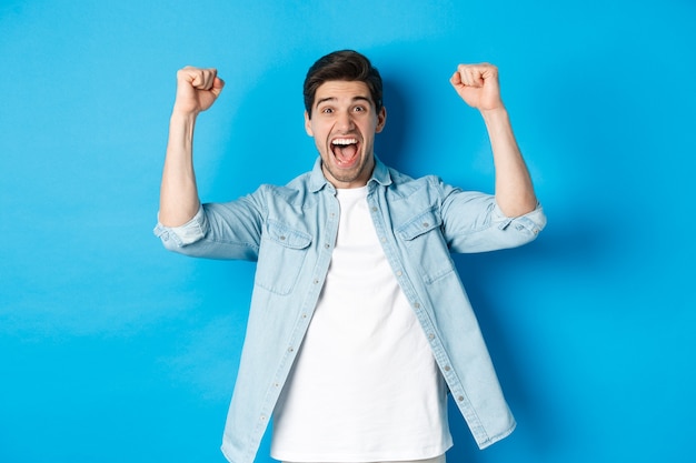 Free photo happy attractive man triumphing, screaming yes and raising hands up to celebrate win, achieve goal, standing against blue background