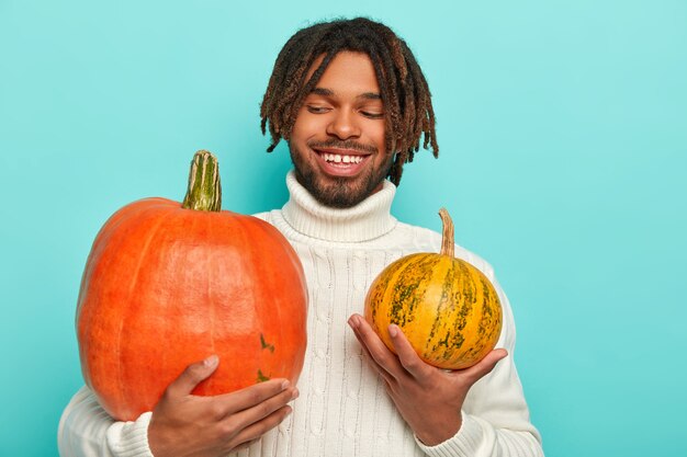 Happy attractive man with pleasant smile, holds big and small pumpkin, chooses product for preapring delicious vegetable cream soup