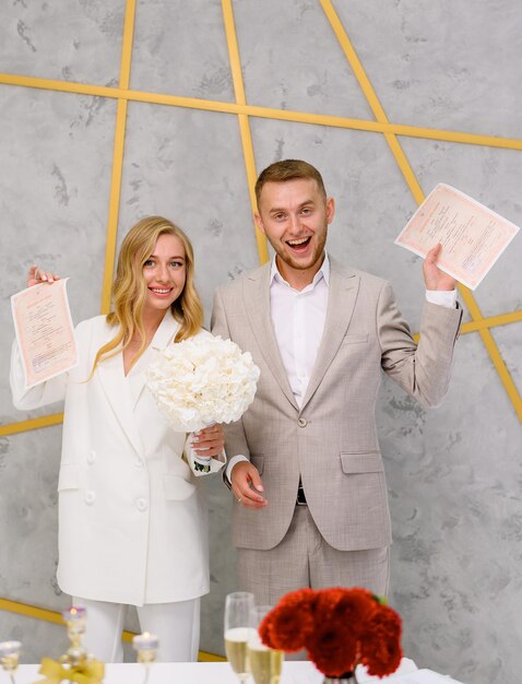 Happy attractive married young man and woman holding wedding license and smiling Wedding day ceremony Cute blonde bride woman wears stylish white wedding suit Family Celebration