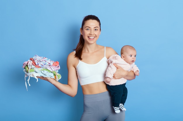 Happy attractive mother with baby isolated over blue background, lady with newborn daughter looks at camera with charming smile, holds stack of kids clothes, takes care of child during maternity leave