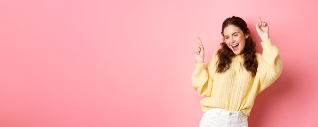 Free photo happy attractive woman having fun pointing fingers up and dancing carefree smiling upbeat standing against pink background