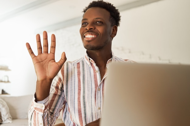 Happy attractive young Afro Amercian male blogger uploading pictures, typing new post for social networks, chatting with his followers online sitting at cafe, waving hand and smiling broadly