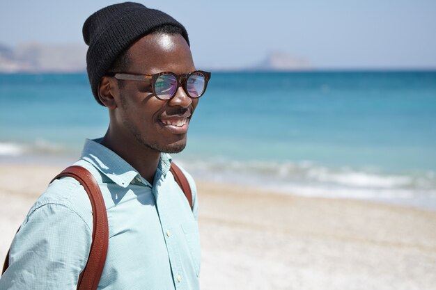 Happy attractive young Afro American man dressed in trendy clothing and accessories relaxing at seaside contemplating azure seascape on calm sunny weather, feeling connection and harmony with nature