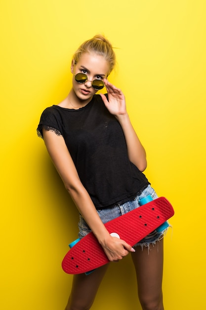 Happy attractive young woman in sunglasses sitting on skateboard over yellow background