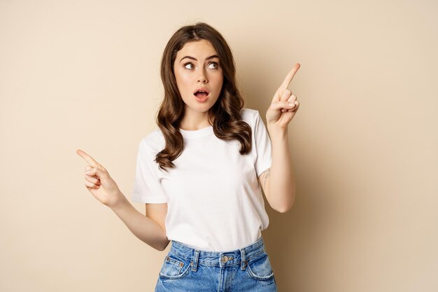 Happy authentic girl smiling, pointing fingers sideways, showing left and right banner, demonstrating promo, standing against beige background