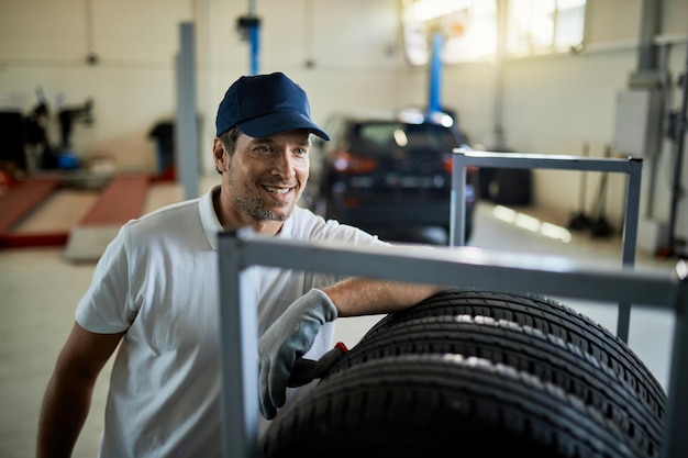 Happy auto mechanic leaning on stack of car tires while working in a workshop