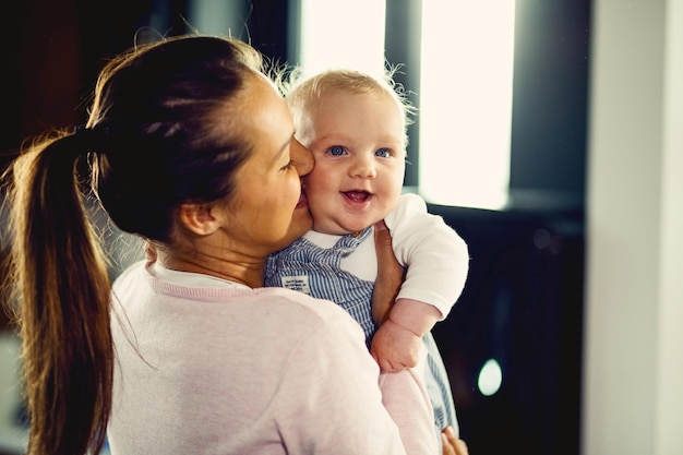 Happy baby boy having fun while mother is kissing him at home