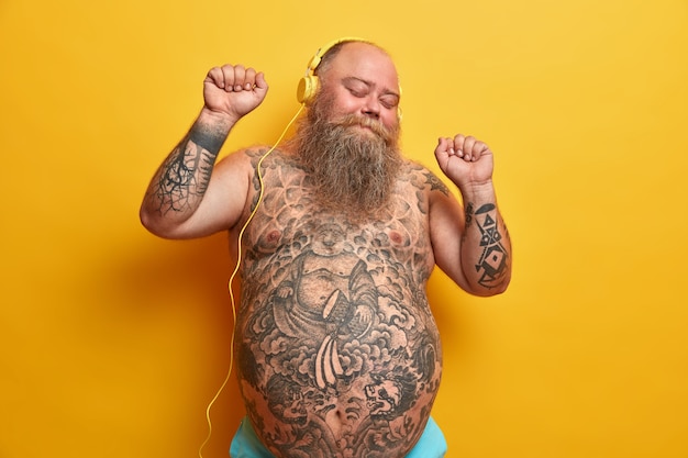 Happy bare man with fat stomach, tattooed belly, enjoys listening new song in headphones, raises arms, clenches fists, moves with rthythm, feels carefree, enjoys fantastic bits, poses indoor