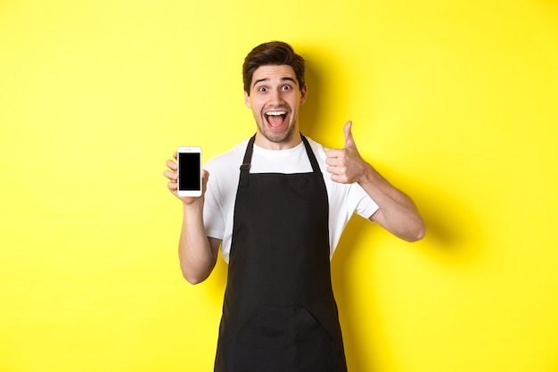 Happy barista in black apron showing smartphone screen, make thumb-up, recommending cafe application, standing over yellow background.