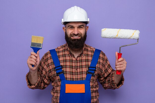 Happy bearded builder man in construction uniform and safety helmet holding paint roller and brush looking smiling cheerfully