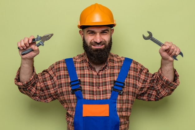 無料写真 建設制服と安全ヘルメットの幸せなひげを生やしたビルダーの男は元気に笑顔に見えるレンチとペンチを保持しています