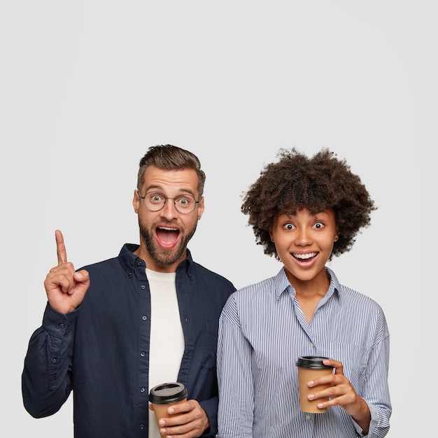 Happy bearded hipster points with index finger upwards, stands near his dark skinned girlfriend