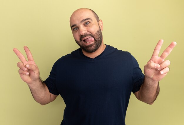 Happy bearded man in black t-shirt  smiling sticking out tongue showing v-sign standing over green wall