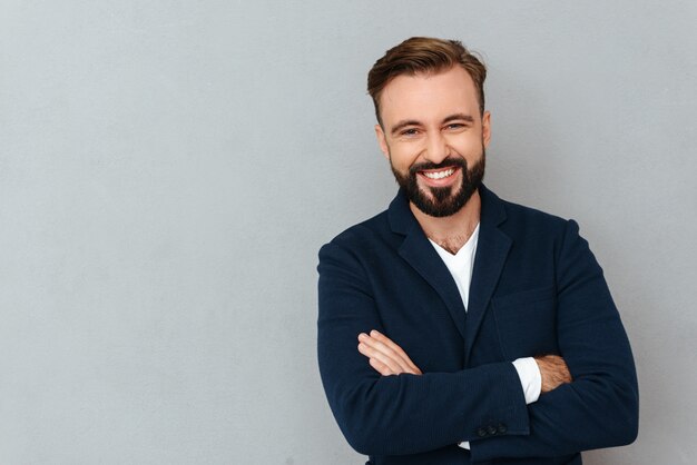 Happy bearded man in busines clothes with crossed arms looking at the camera over gray 