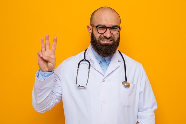 Happy bearded man doctor in white coat with stethoscope around neck wearing glasses looking smiling showing number three with fingers