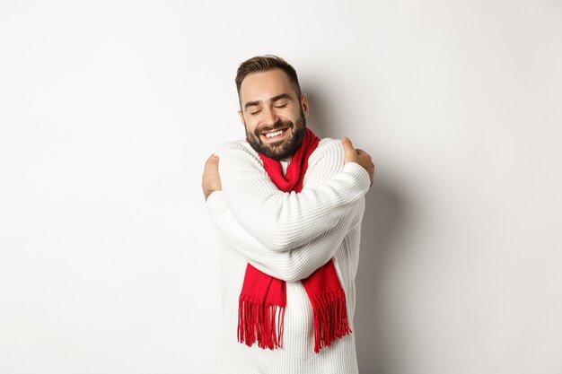 Happy bearded man enjoying comfortable winter sweater, smiling with warmth, hugging himself, standing over white background