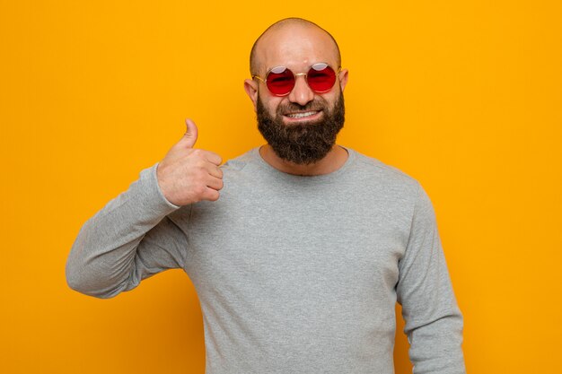 Happy bearded man in grey sweatshirt wearing red glasses looking smiling showing thumb up