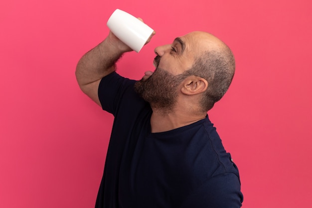 ピンクの壁の上に立って飲むためにお茶を持っている海軍のTシャツで幸せなひげを生やした男