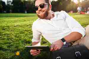 Free photo happy bearded man in sunglasses lying on grass outdoors with tablet computer and looking away