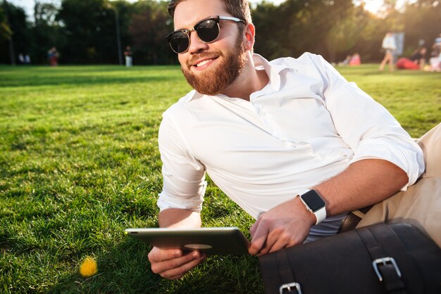 タブレットコンピューターで屋外の芝生に横になっているとよそ見サングラスで幸せなひげを生やした男