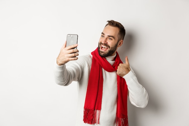 Happy bearded man video call and showing thumbs up at mobile phone, like christmas gift, talking online, standing over white background