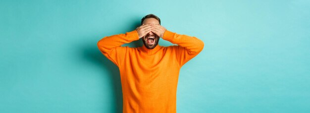 Happy bearded man waiting for surprise shut ears and expeting gifts standing over light blue backgro