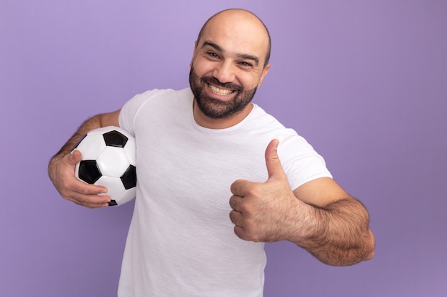 Happy bearded man in white t-shirt holding soccer ball  smiling cheerfully showing thumbs up standing over purple wall