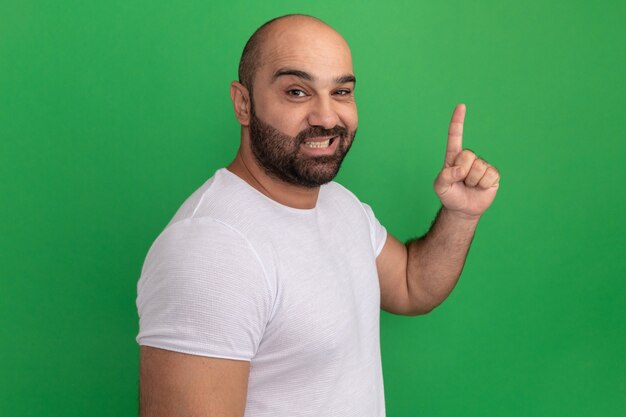 녹색 벽 위에 서 검지 손가락을 보여주는 웃 고 흰색 티셔츠에 행복 한 수염 난된 남자