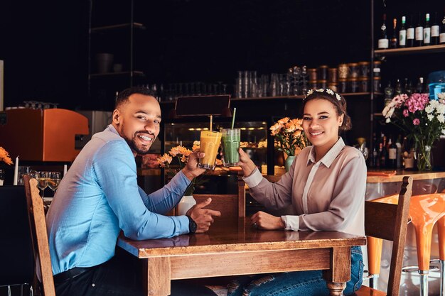 Happy beautiful African-American couple in love having a great time together in a restaurant at their dating, an attractive couple enjoying each other, holds glasses of a juice.