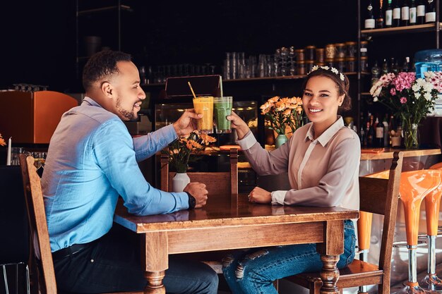 Happy beautiful African-American couple in love having a great time together in a restaurant at their dating, an attractive couple enjoying each other, holds glasses of a juice.