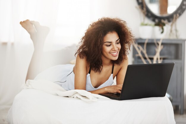 ノートパソコンを見て笑みを浮かべて自宅のベッドに横になっているパジャマで幸せなアフリカ美女。