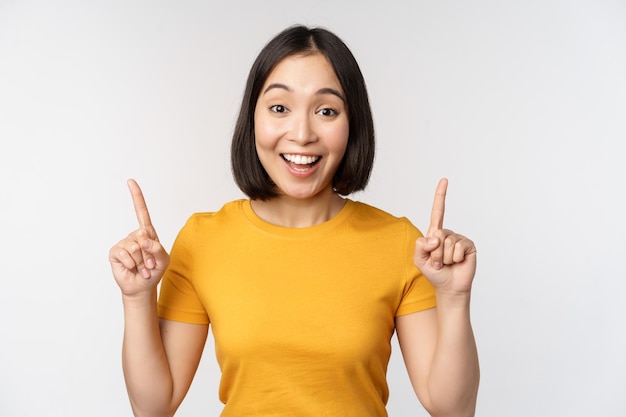 Happy beautiful asian girl showing advertisement pointing fingers up standing over white background