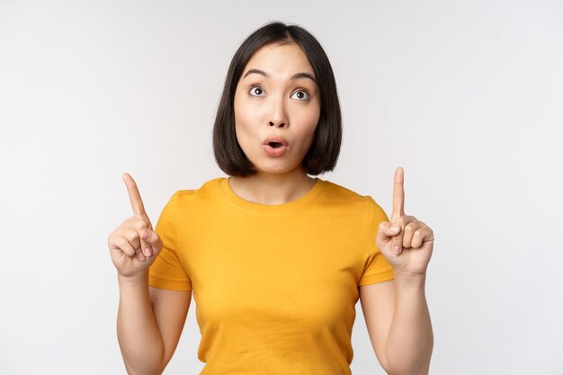 Happy beautiful asian girl showing advertisement pointing fingers up standing over white background