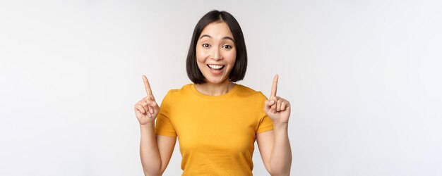 Happy beautiful asian girl showing advertisement pointing fingers up standing over white background