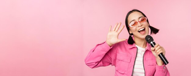 Happy beautiful asian girl singing with mic using microphone enjoying karaoke posing against pink studio background