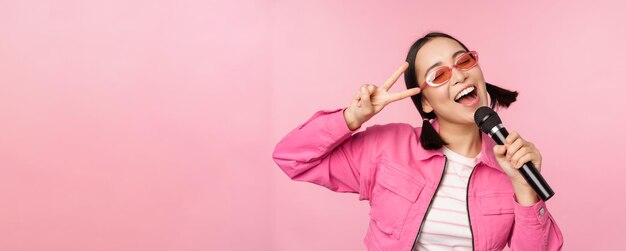 핑크색 스튜디오 배경에서 포즈를 취하는 노래방을 즐기고 있는 마이크를 사용하여 마이크를 들고 노래하는 행복한 아름다운 아시아 소녀