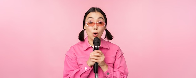 Happy beautiful asian girl singing with mic using microphone enjoying karaoke posing against pink studio background