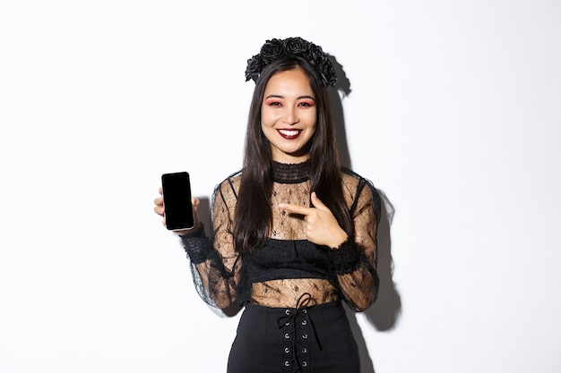 Happy beautiful asian girl in witch costume pointing finger at smartphone screen with pleased smile, showing halloween announcement, white background.