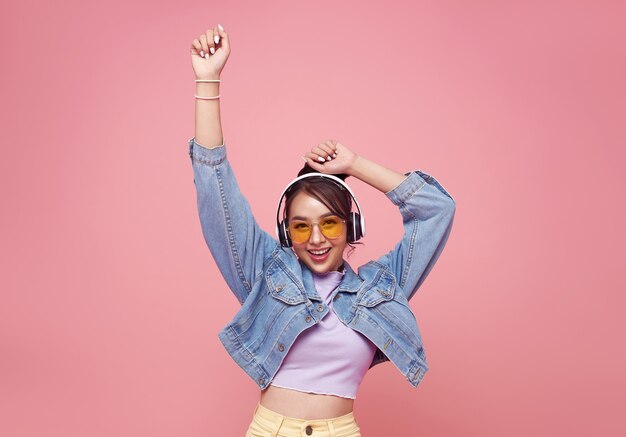 Happy beautiful Asian teen woman in yellow glasses listening music in headphones and dance on pink wall.