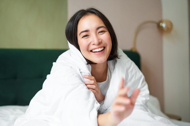 Happy beautiful asian woman covered with blanket sits on bed in warm duvet tries to reach camera wit