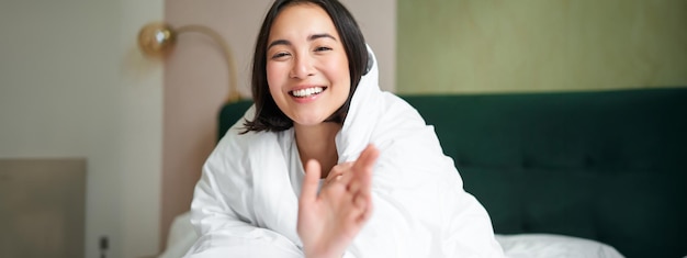 毛布で覆われた幸せな美しいアジアの女性が暖かい羽毛布団のベッドに座ってカメラに到達しようとします