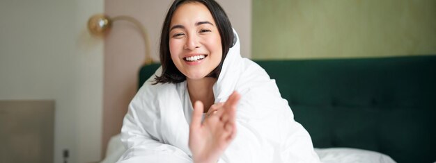 happy beautiful asian woman covered with blanket sits on bed in warm duvet tries to reach camera