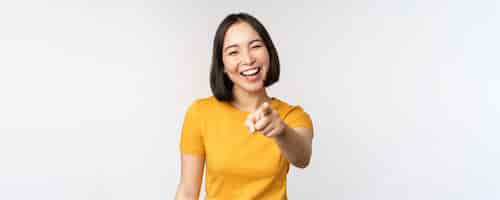 Free photo happy beautiful asian woman laughing pointing finger at camera and chuckle smiling carefree standing in yellow tshirt over white background