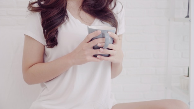 Happy beautiful Asian woman smiling and drinking a cup of coffee or tea on bed