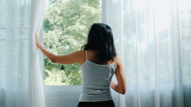 Happy beautiful Asian woman smiling and drinking a cup of coffee or tea near the window in bedroom. Young latin girl open curtains and relax in morning. Lifestyle lady at home .