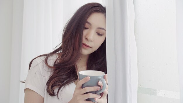 幸せと美しいアジアの女性の笑みを浮かべて、寝室の窓の近くのコーヒーや紅茶のカップを飲みます。
