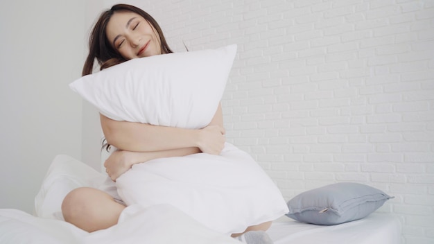 幸せな美しいアジアの女性は目を覚ます、笑みを浮かべて、寝室の彼女のベッドで彼女の腕を伸ばします。