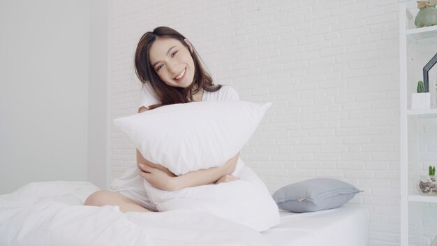 행복 한 아름 다운 아시아 여자 일어나, 웃 고 침실에서 그녀의 침대에서 그녀의 팔을 스트레칭.